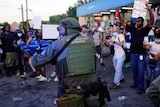 A man dressed in SWAT uniform wearing a helmet and bulletproof vest points a gun off camera in front of protesters