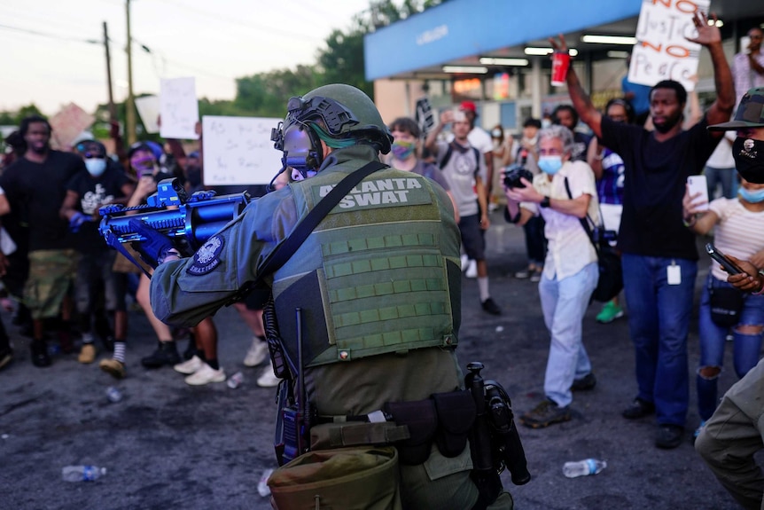 A man dressed in SWAT uniform wearing a helmet and bulletproof vest points a gun off camera in front of protesters