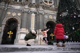 A little girl and her family wearing puffy coats jumps in the air outside a large christmas tree in front of a church