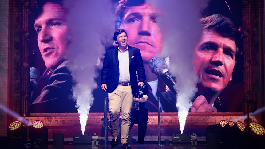 Tucker Carlson walks through smoke machines, behind three large images of himself