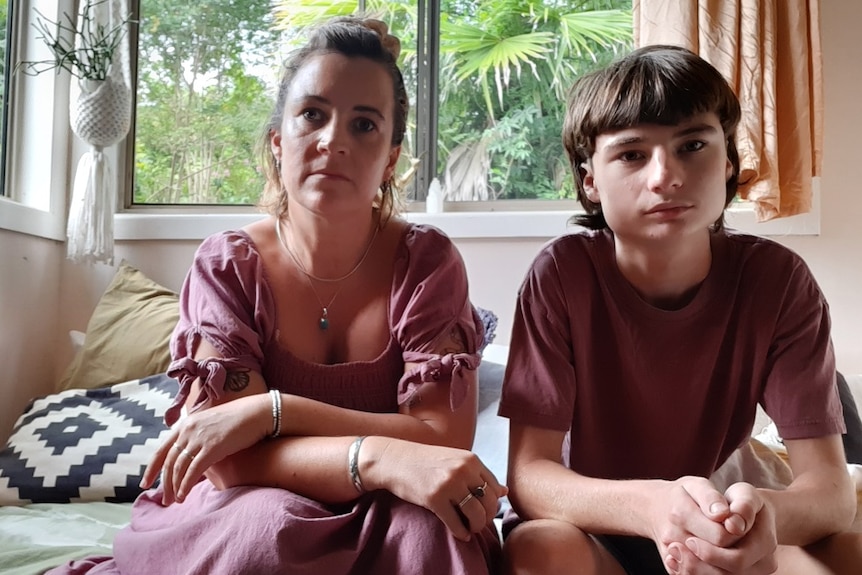 A woman and her teenage son sit on a bed, stare unsmiling at the camera