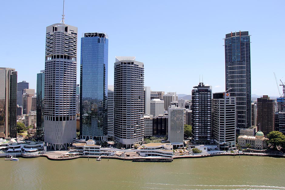 Tall office buildings line the bank of a brown river.