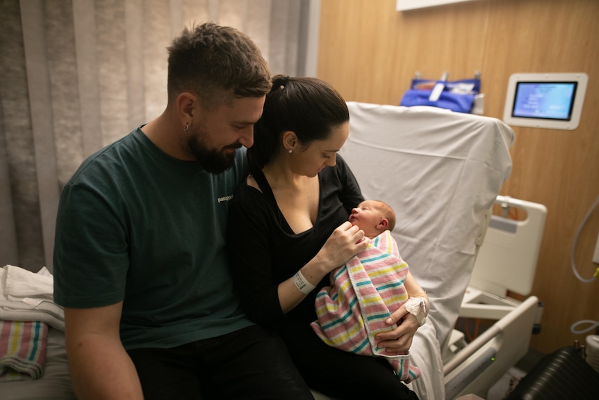 Taryn and Nathan sit together on a maternity bed holding their newborn baby.