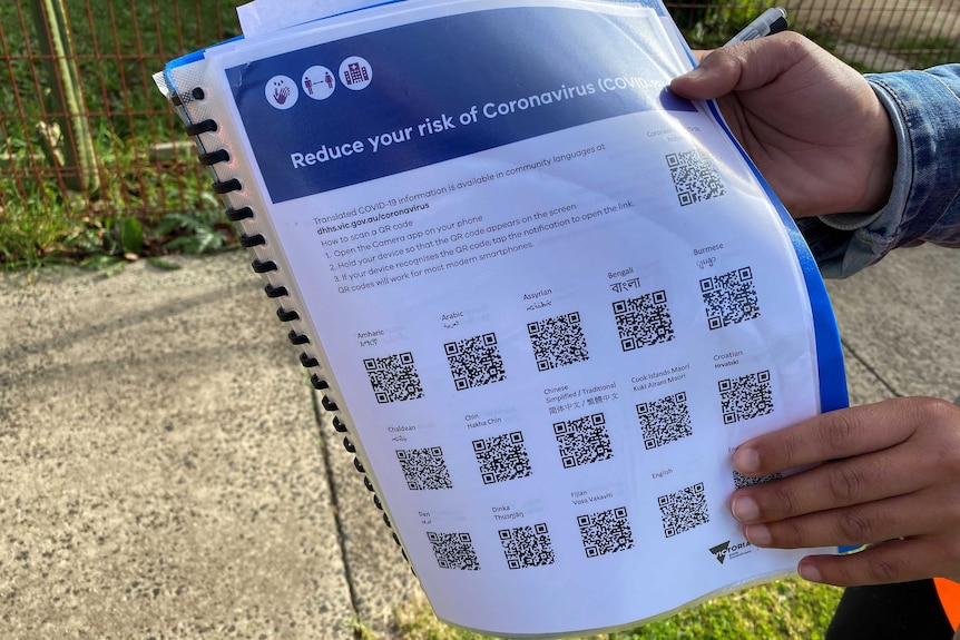 A person holds a document titled "Reduce your risk of Coronavirus" with QR codes for 15 different languages.