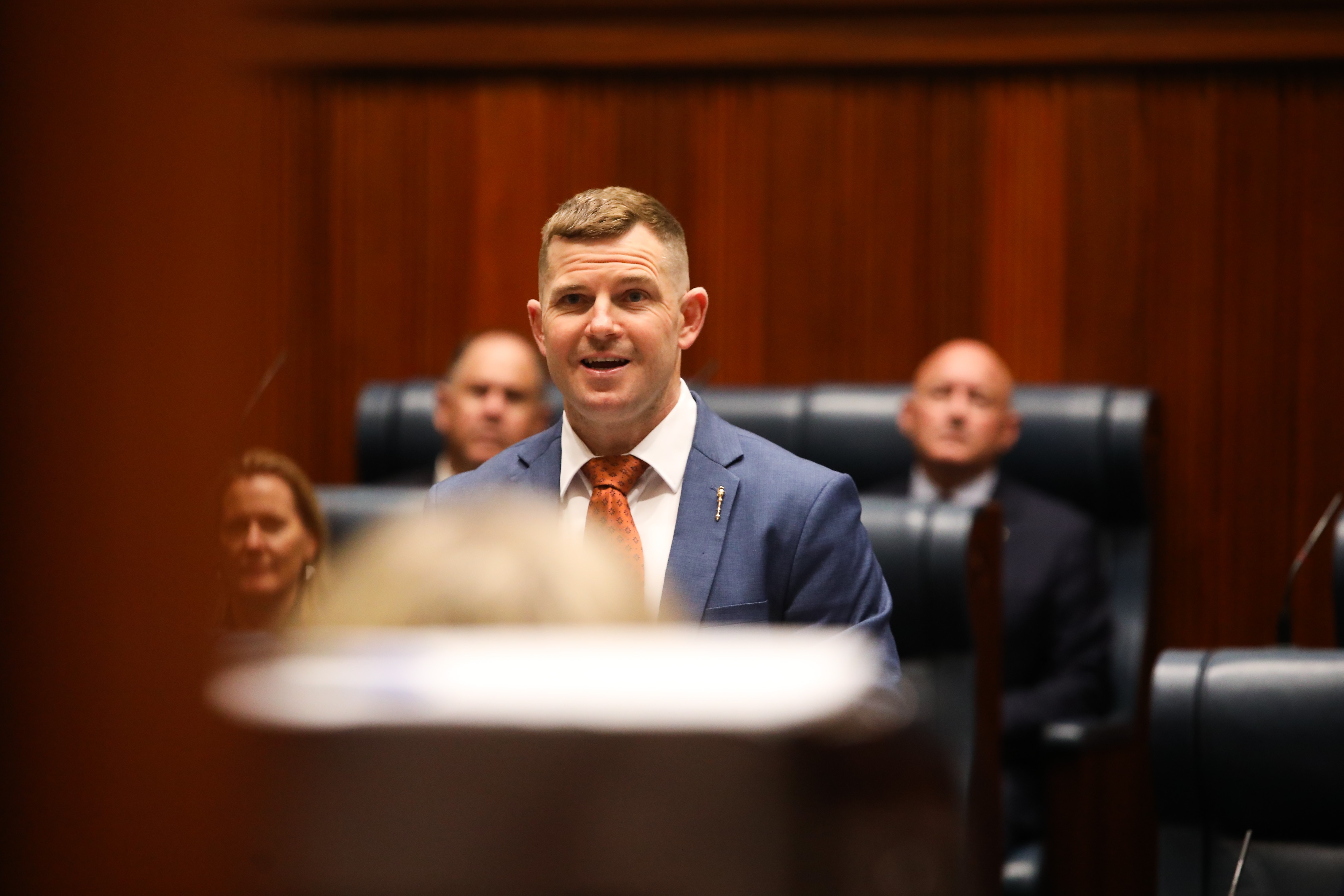 An MP speaks in Parliament, with other MPs seated behind him, blurred.