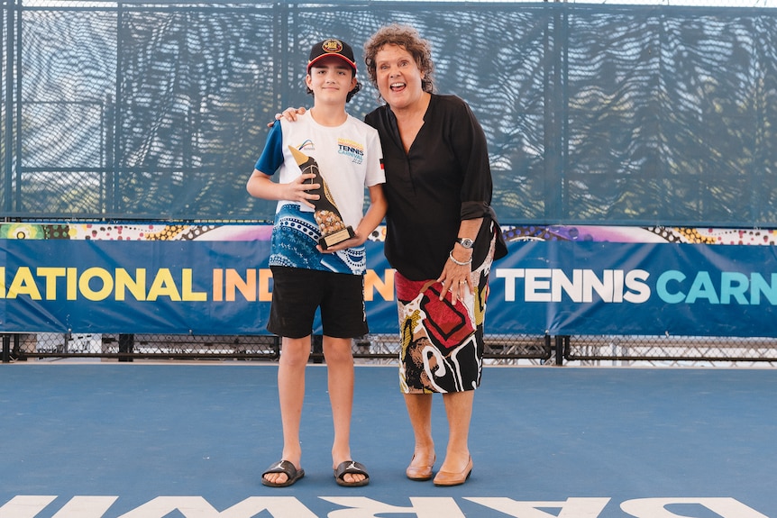 An Aboriginal kid holds a trophy and stands next to an older Aboriginal woman on a tennis court in Darwin.