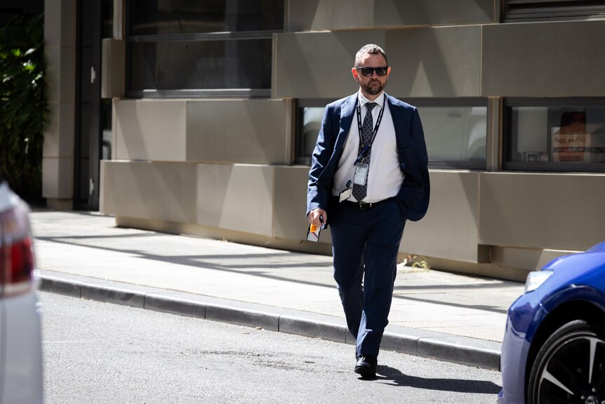A man wearing sunglasses and a dark blue suit and a grey tie walking forwards across a road.