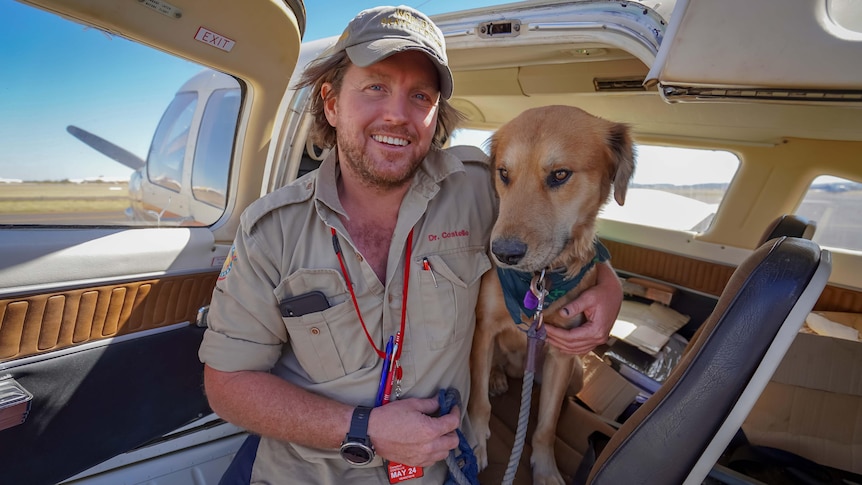 A man has his arm around a dog. They both stand in front of a small plane. 