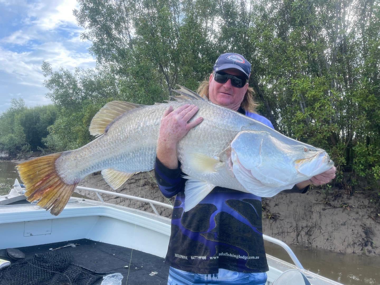 Un hombre con sombrero y gafas de sol sostiene un barramundi gigante