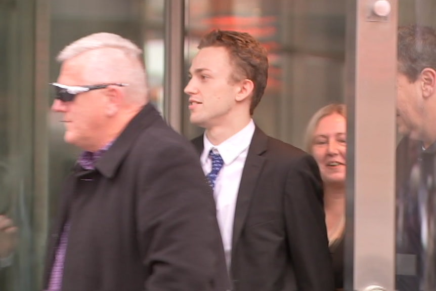 A young man in a suit and tie leaves the glass doors of court.
