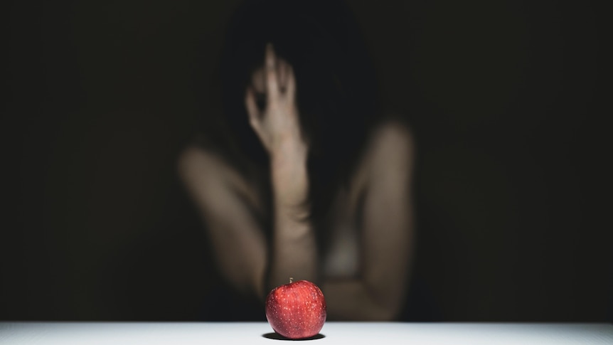 A blurry distraught women clutches her face with her hand. In front of her is a red apple.
