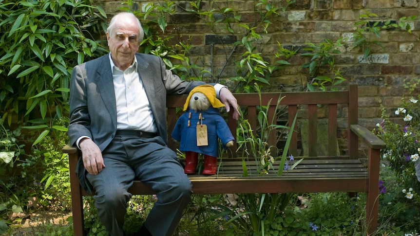 British author Michael Bond sits on a bench in a garden next to a Paddington Bear toy.