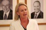 Fiona Nash standing in front of two black and white portraits of Nationals leaders. She is wearing a white blazer.