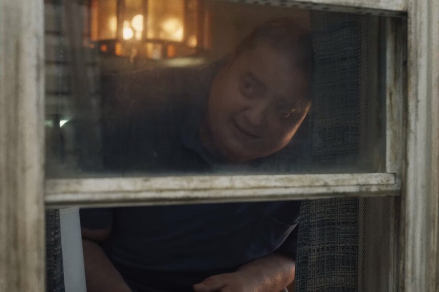 White middle-aged, overweight and balding man with blue eyes peers through a window from the inside of a darkened house.