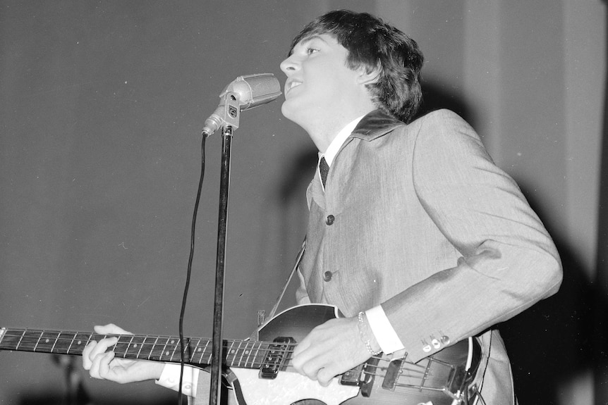 Paul McCartney singing on stage at a microphone, wearing a suit, holding a bass.