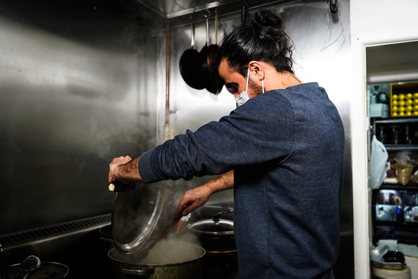A man wearing a mask, stirs a steaming pot of food in an industrial kitchen.