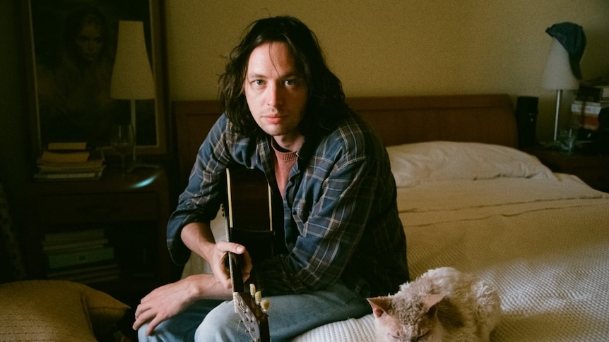 Nick Weaver sits on a bed, holding a guitar, next to a cat, looking at the camera