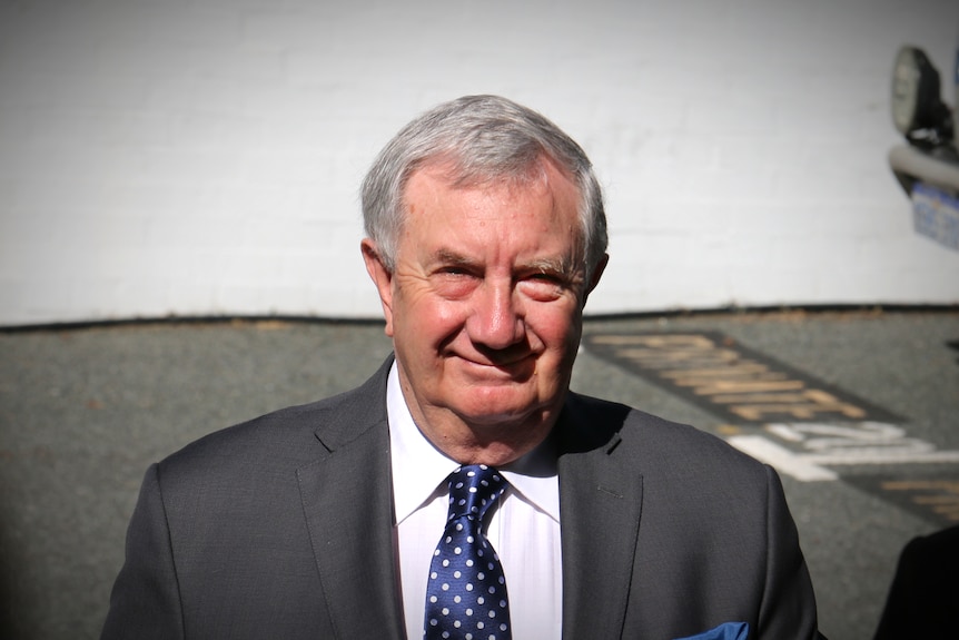 A man with greying hair and a grey suit walks in a car park