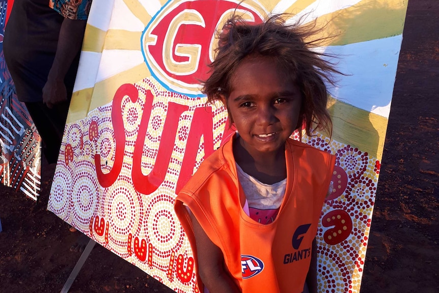 A young child stands in front of a bonnet painted with an AFL team's logo.