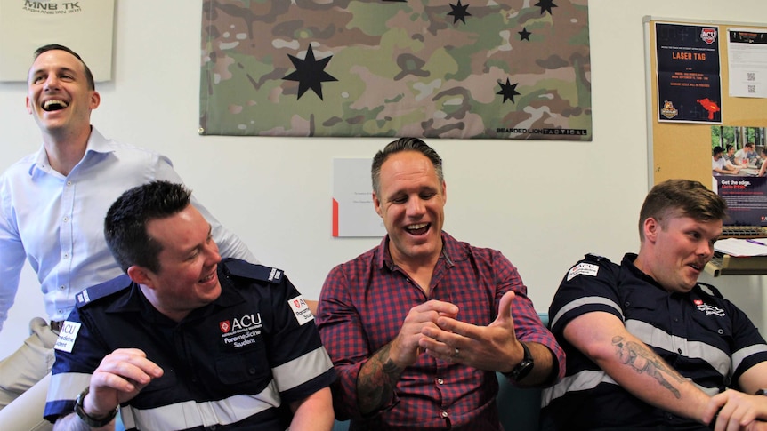 Four men laugh together in front of a Australian flag printed on camouflage fabric, two in student paramedic uniforms.