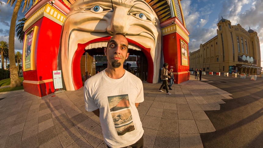 a man wearing a white shirt stands in front of Luna Park in Saint Kilda