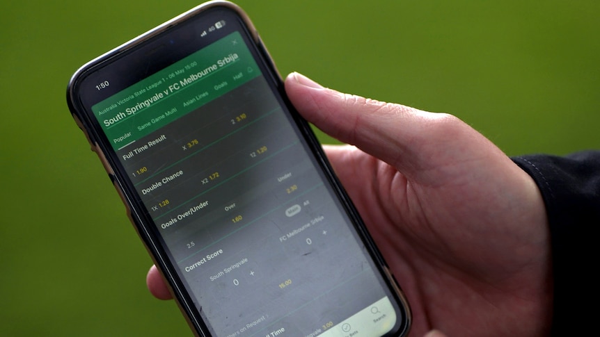 A hand holds a mobile phone showing a betting app. The screen shows the local soccer game is available to bet on.
