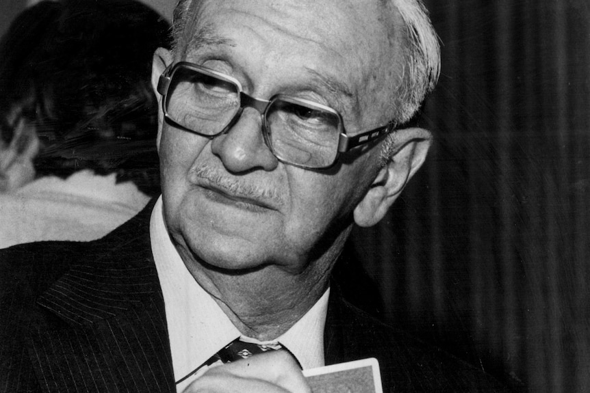 An older man with thick glasses and grey hair holding a handful of cards.