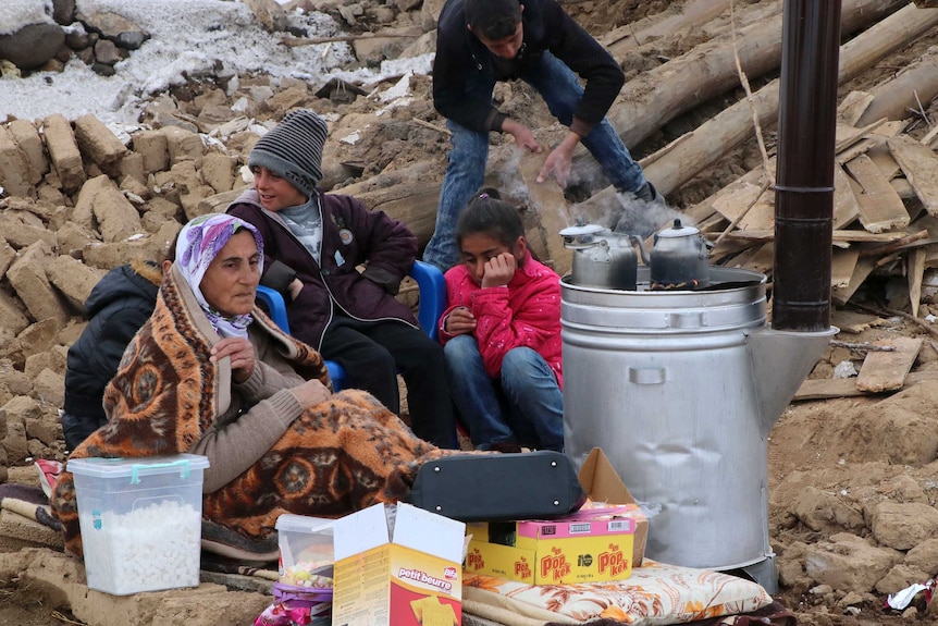 An elderly woman wearing a head scarf sits on the ground with boy and girl next to rocks and snow.