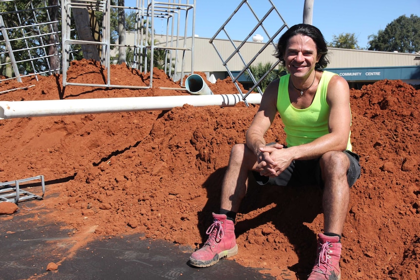 A man sits on a pile of red dirt in front of miscellaneous metal items and pipes