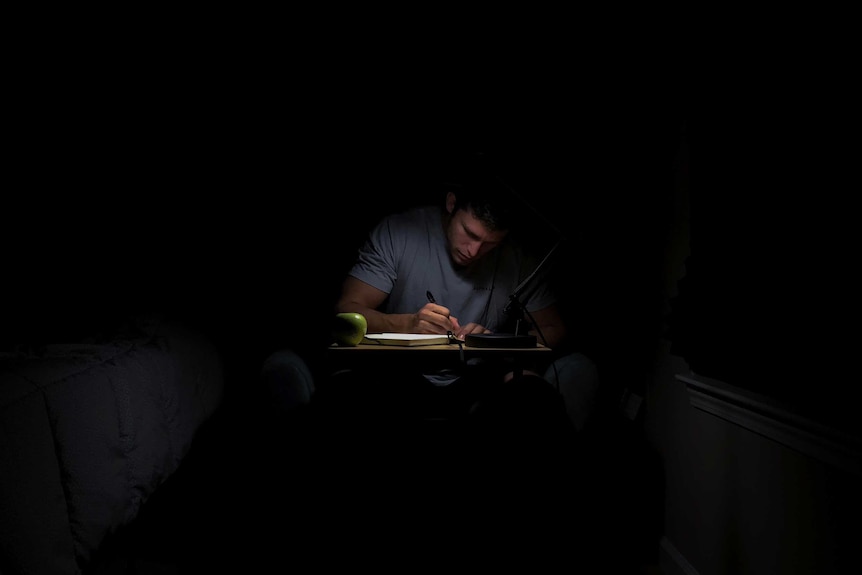 A student studying for university at night in a dark room, a desk lamp is lighting only his face and his notes.