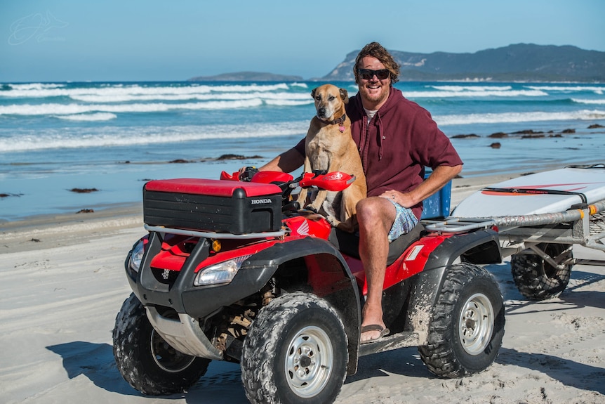 Luc Longley on a quad bike on the sand at a beach with his dog on the seat in front of luc