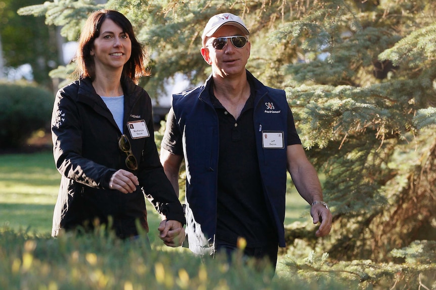 MacKenzie and Jeff Bezos walk by pine trees, wearing name tags and dressed casual clothing.