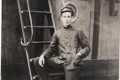 A young man poses from the camera on a ship wearing a sailors uniform. black and white