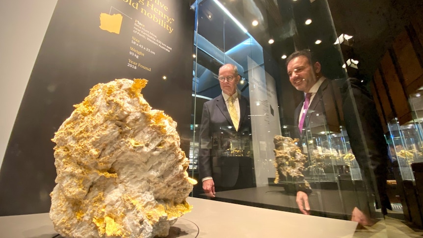 Two men in suits looking through a glass window at a rare gold specimen.   
