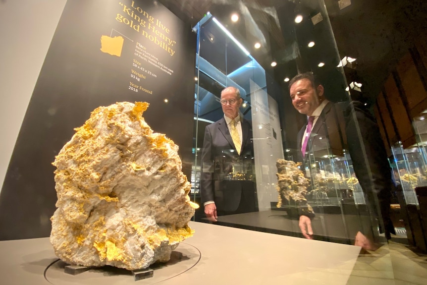 Two men in suits looking through a glass window at a rare gold specimen.   