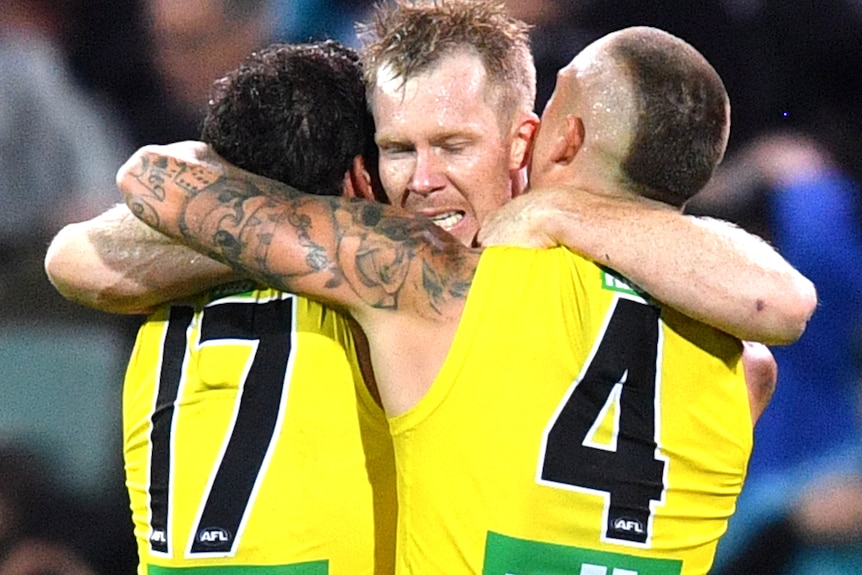Jack Riewoldt's face can be seen as he hugs Daniel Rioli and Dustin Martin, whose backs are to camera