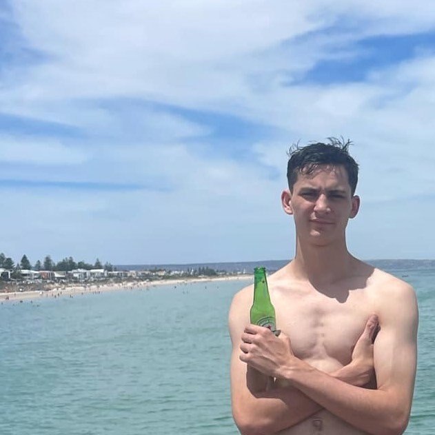 Rylie Lewis Everson-Clare holds a beer bottle in his hand with a beach behind him