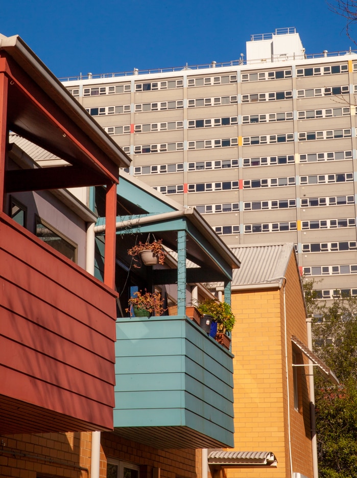 On a bright blue day, you see three colourful gable-roofed houses set against a large concrete public housing tower.