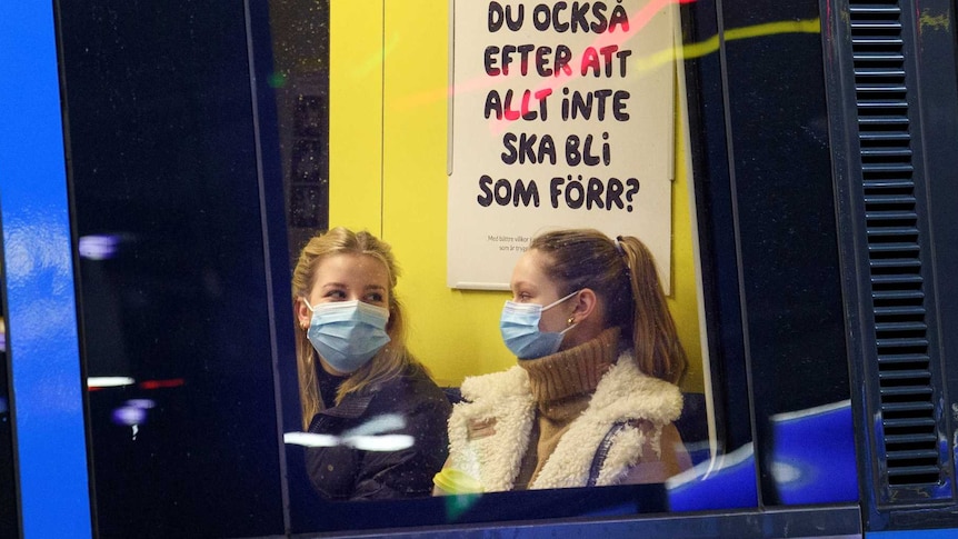 two women in masks look at each other as they chat on a train
