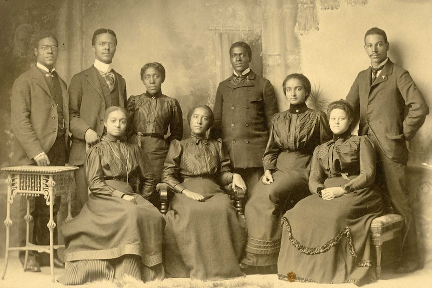 A black and white photo of nine people in formal dress, curtains in background.