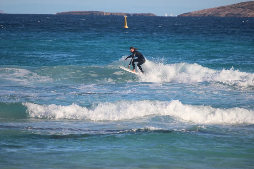 A man surfing a wave in a wetsuit. A yellow satellite receiver is visible in the water behind him.
