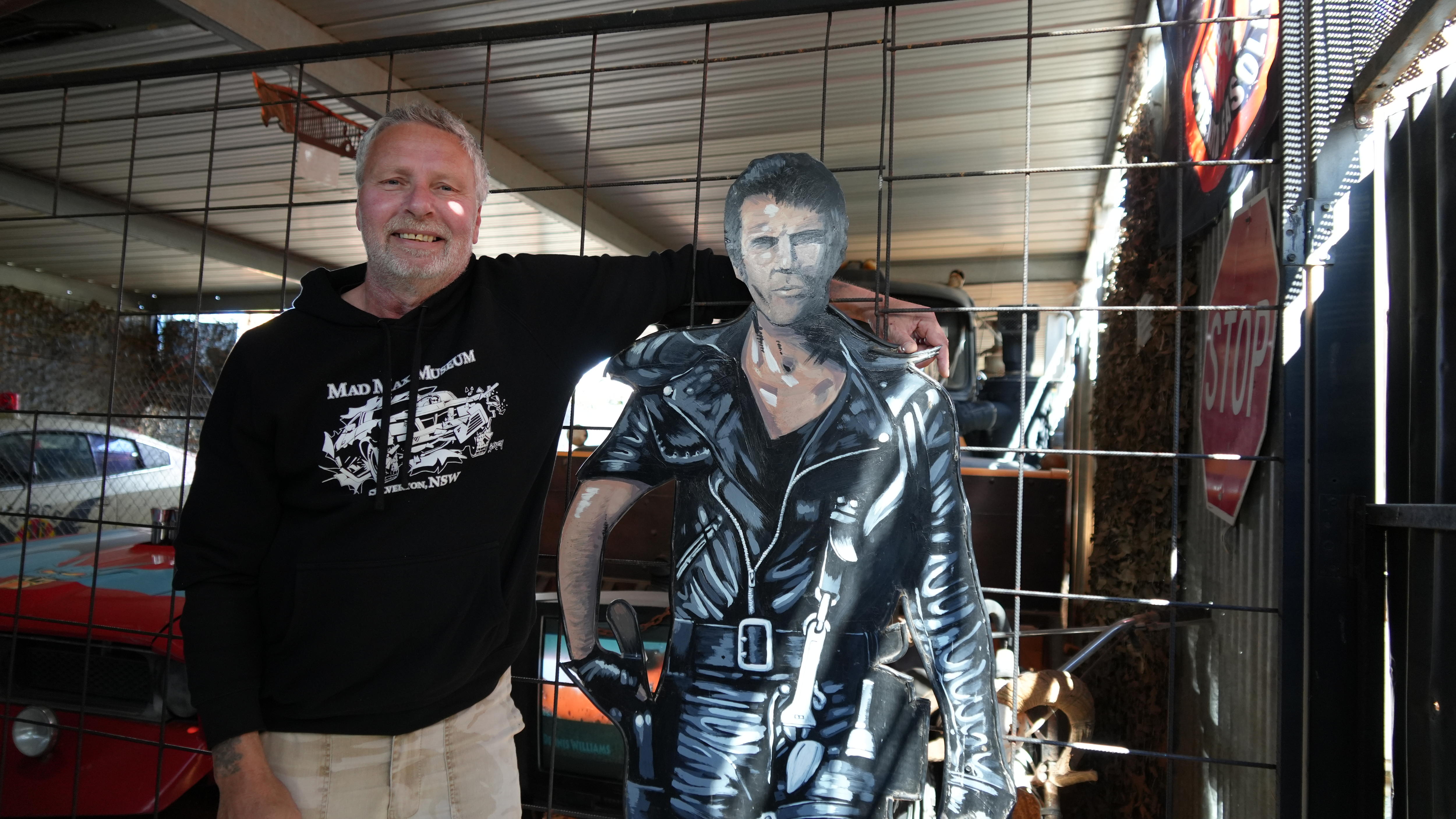 A middle aged man in a black hoodie standing in front under cover with his arm around a cut out of a man wearing bike leathers.
