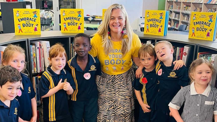 A woman in the middle of 7 primary school students with book shelves behind them. 