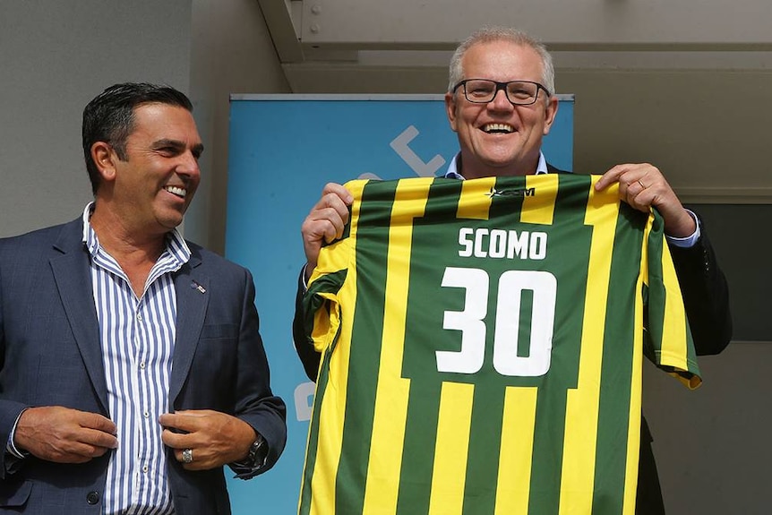 Two men smile, one holds up a green and yellow soccer shirt