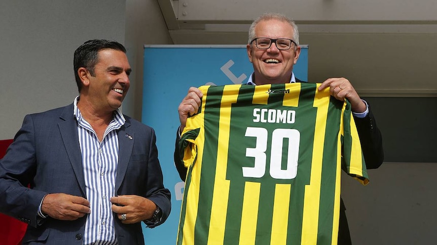 Two men smile, one holds up a green and yellow soccer shirt