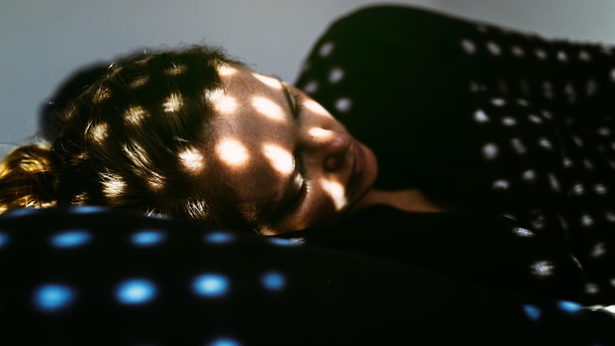 A woman lies on a bed in sunlight dappled by shadows. She looks concerned.