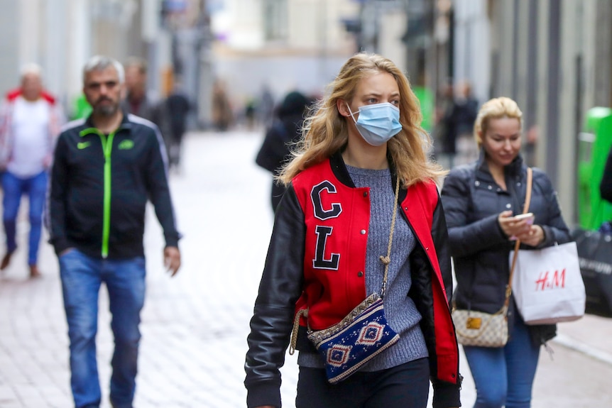 A young woman with long blonde hair wears a blue surgical mask on an Amsterdam street