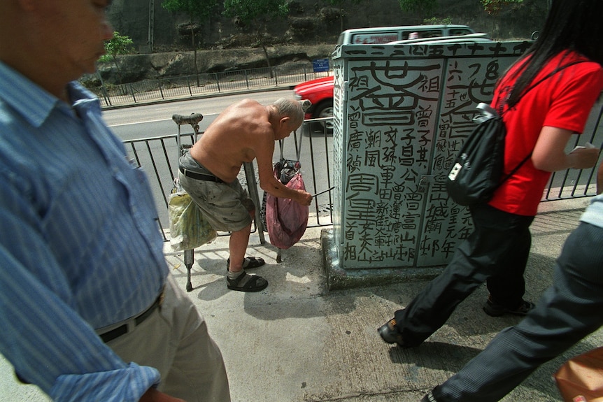 An old man bent over painting black calligraphy with a paint brush on a public box.