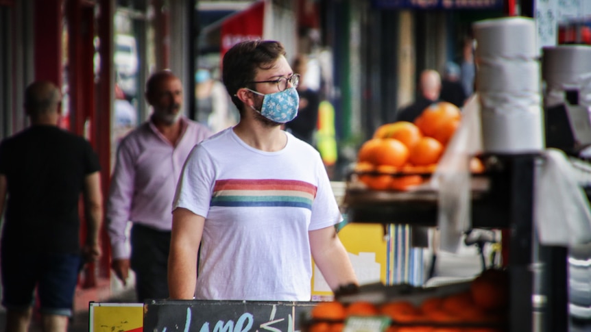 A person in a white shirt with a rainbow stripe and a blue cloth mask shops at a streetside grocer.