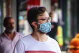 A person in a white shirt with a rainbow stripe and a blue cloth mask shops at a streetside grocer.